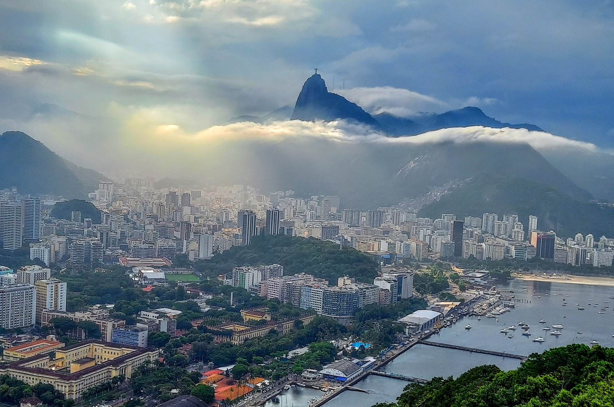 The Christ the Redeemer statue on top of dramatic Corcovado mountain shrouded by clouds, whose beams of light shine onto the vibrant city scape of Rio de Janeiro, Brazil.